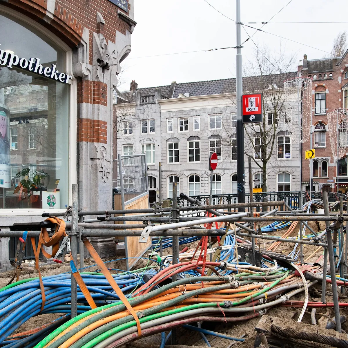 A dug-up street in Amsterdam with bundles of blue, green, orange and red cables and fibre ducts, under scaffolding tubes